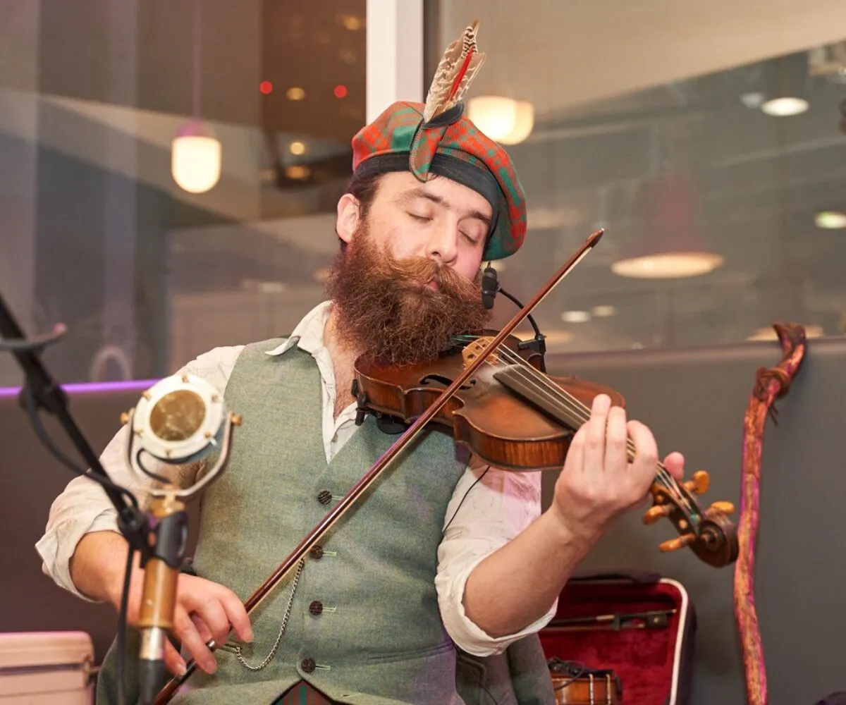 Scottish fiddler in traditional attire performing live at a curated brand activation event by The Curators of Exiled Scots. Dressed in tartan and feathered bonnet, the artist delivers an authentic musical experience blending heritage and storytelling, tailored for luxury clients, cultural partners, and government audiences seeking high-quality Scottish entertainment with global impact.