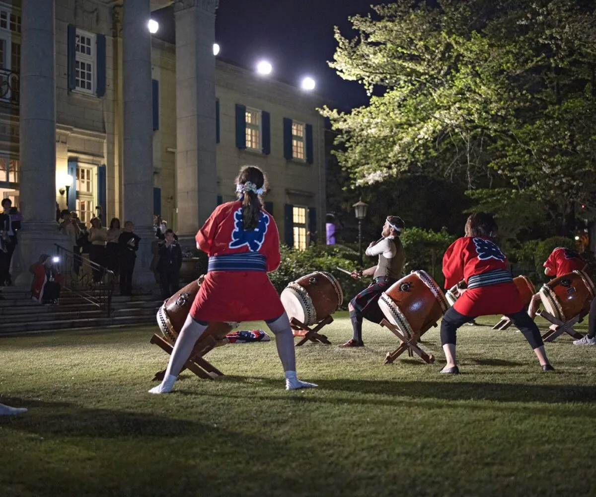 Outdoor evening performance featuring a collaboration between Japanese Taiko drummers and a Scottish Highland percussionist in traditional kilt, staged on the lawn of a grand diplomatic residence. Curated by The Curators of Exiled Scots